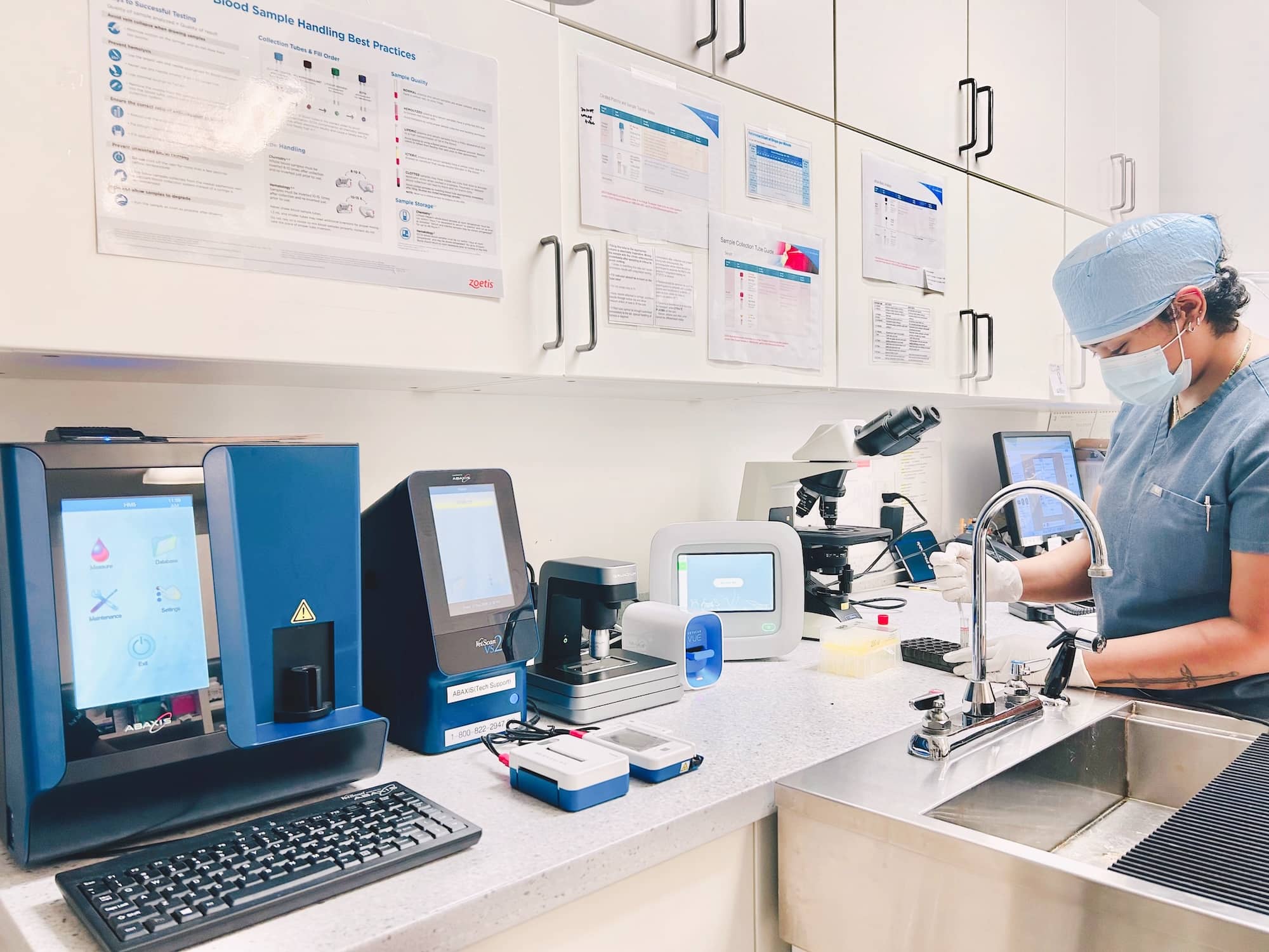 A veterinarian in surgical attire working in a laboratory, with various diagnostic equipment and a microscope on the countertop, and informational posters on the wall above.